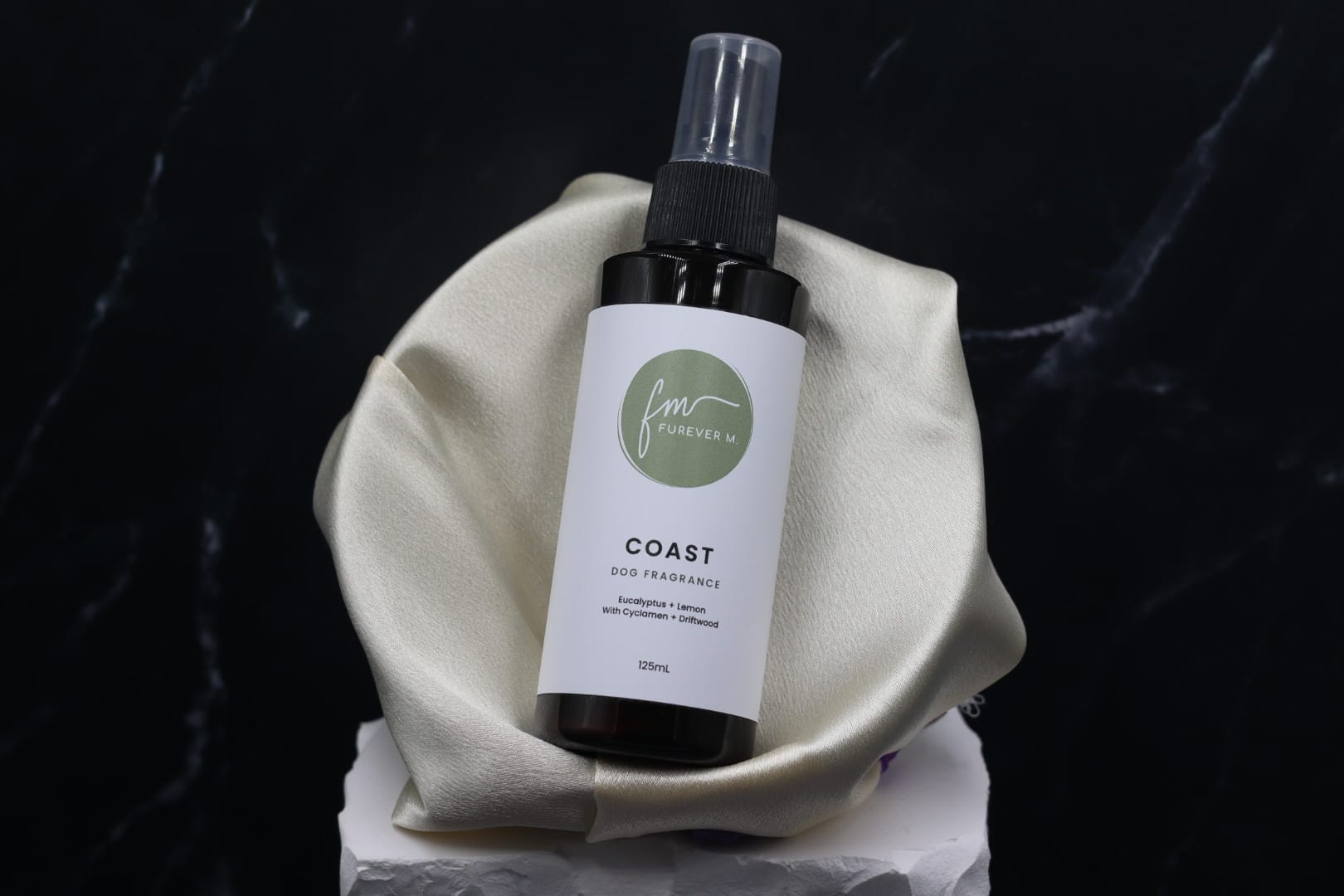 A black spray bottle of the Coast dog cologne with it's white label with the green Furever M logo on it, placed in a silken bowl against a black marble backdrop.