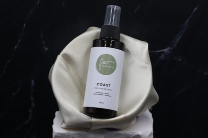 A black spray bottle of the Coast dog cologne with it's white label with the green Furever M logo on it, placed in a silken bowl against a black marble backdrop.