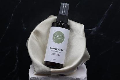 A black spray bottle of the Wilderness dog cologne with it's white label with the green Furever M logo on it, placed in a silken bowl against a black marble backdrop.