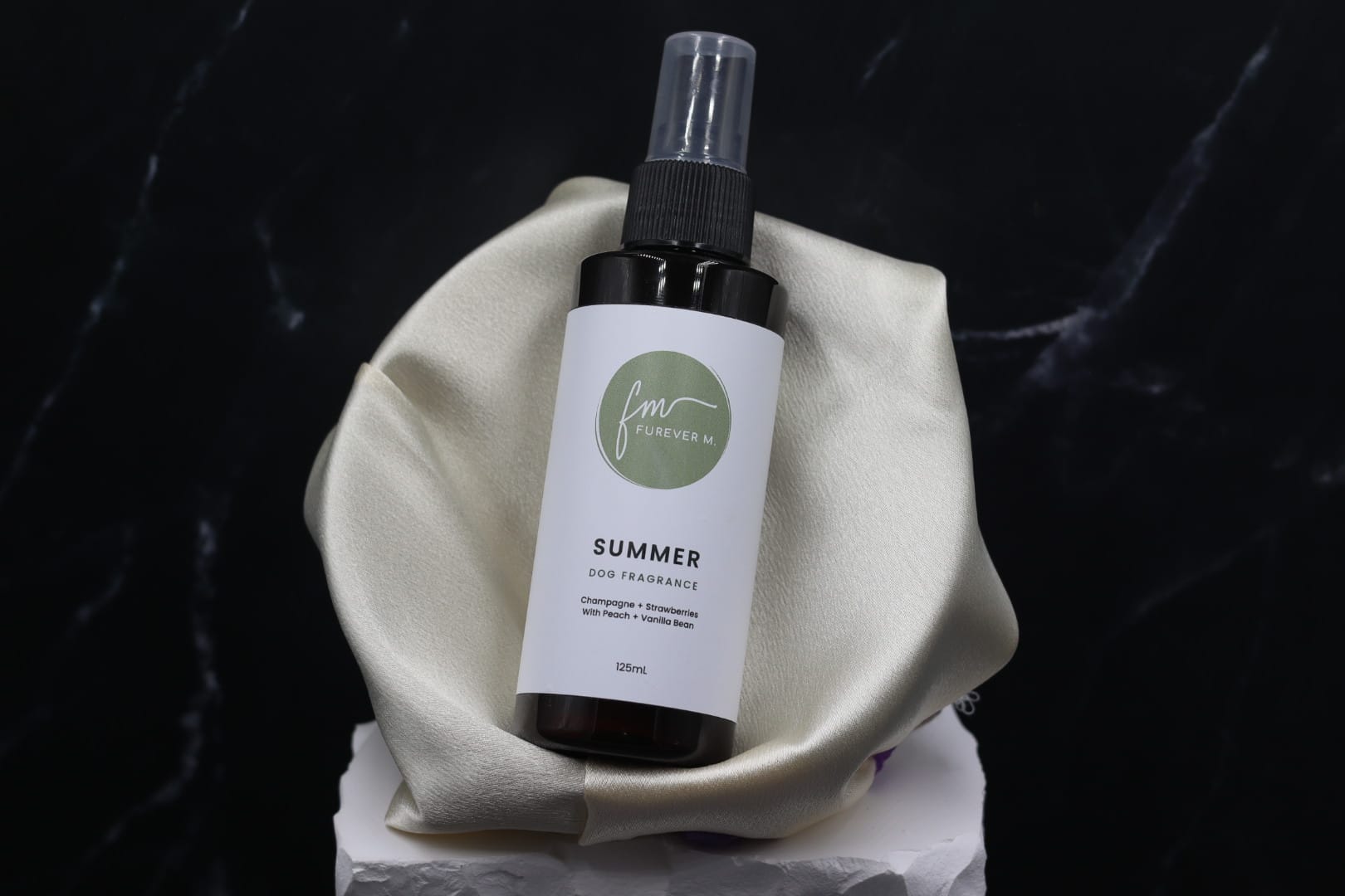 A black spray bottle of the Summer dog cologne with it's white label with the green Furever M logo on it, placed in a silken bowl against a black marble backdrop.