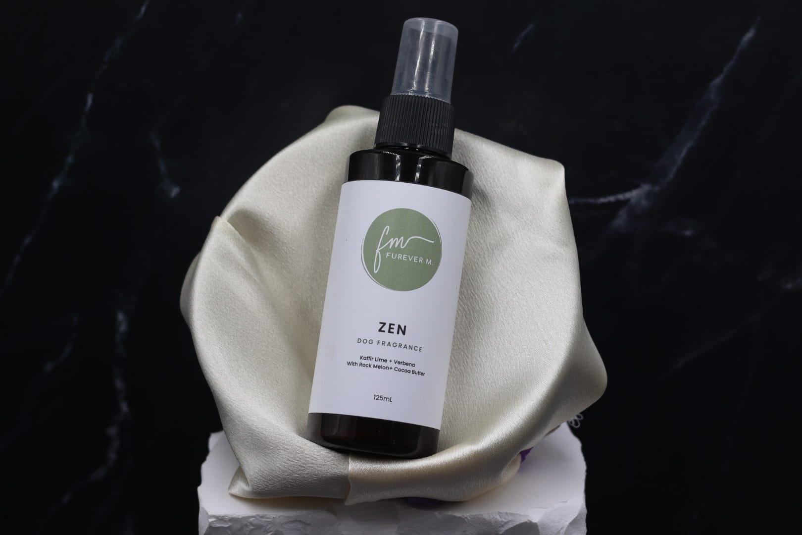 A black spray bottle of the Zen dog cologne with it's white label with the green Furever M logo on it, placed in a silken bowl against a black marble backdrop.