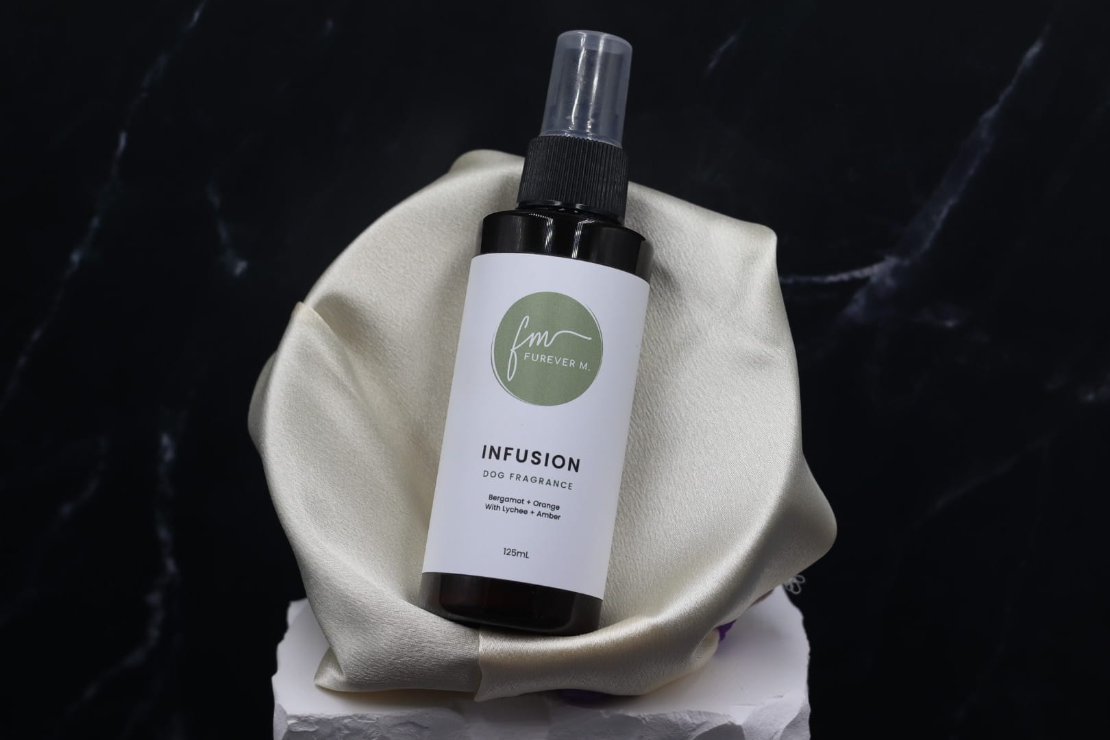 A black spray bottle of the Infusion dog cologne with it's white label with the green Furever M logo on it, placed in a silken bowl atop a black marble backdrop.