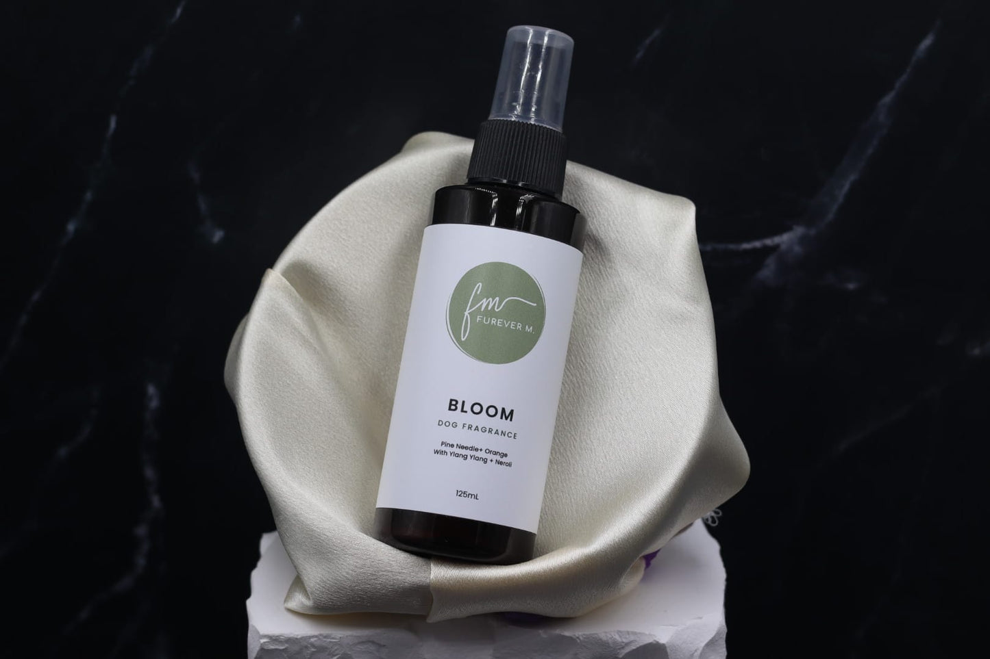 A black spray bottle of the Bloom dog cologne with it's white label with the green Furever M logo on it, placed in a silken bowl against a black marble backdrop.