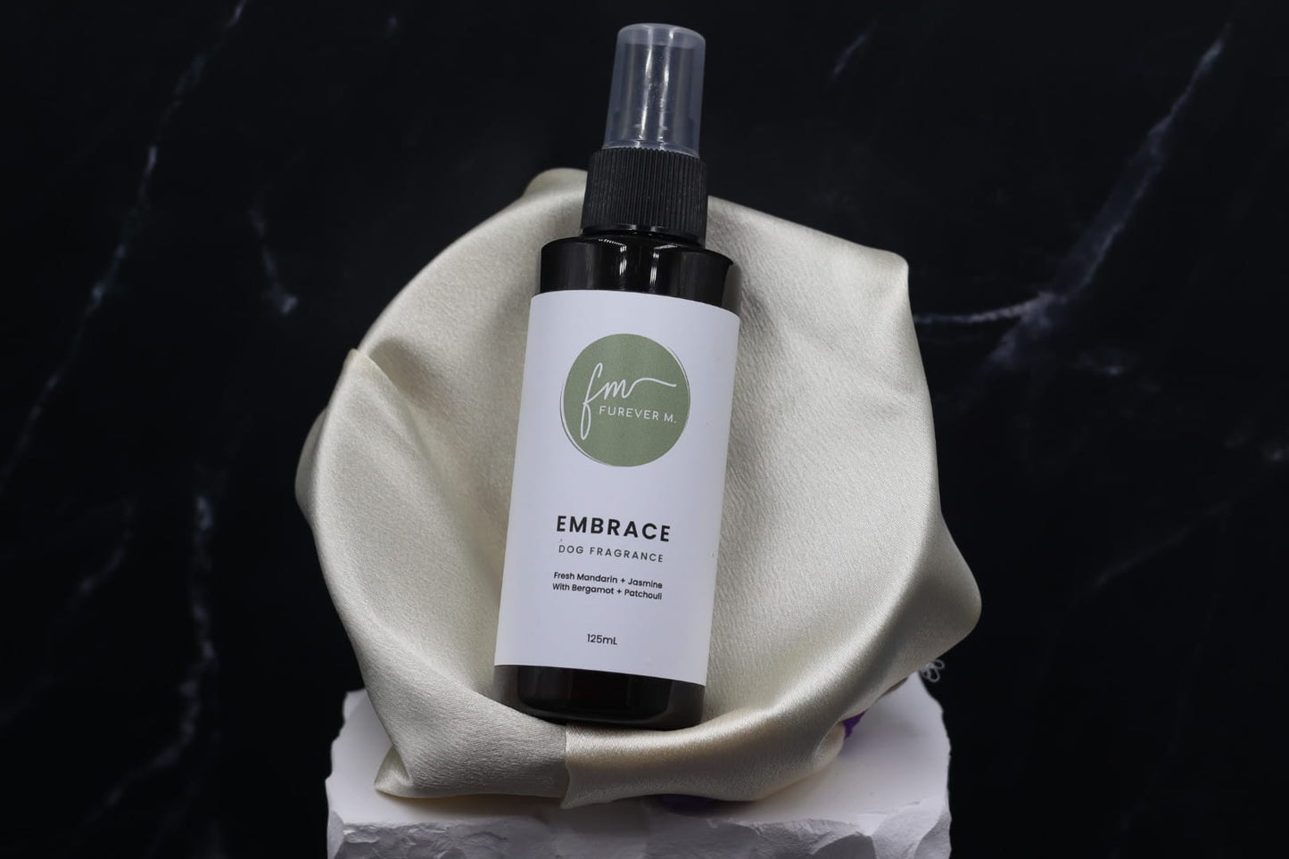 A black spray bottle of the Embrace dog cologne with it's white label with the green Furever M logo on it, placed in a silken bowl against a black marble backdrop.