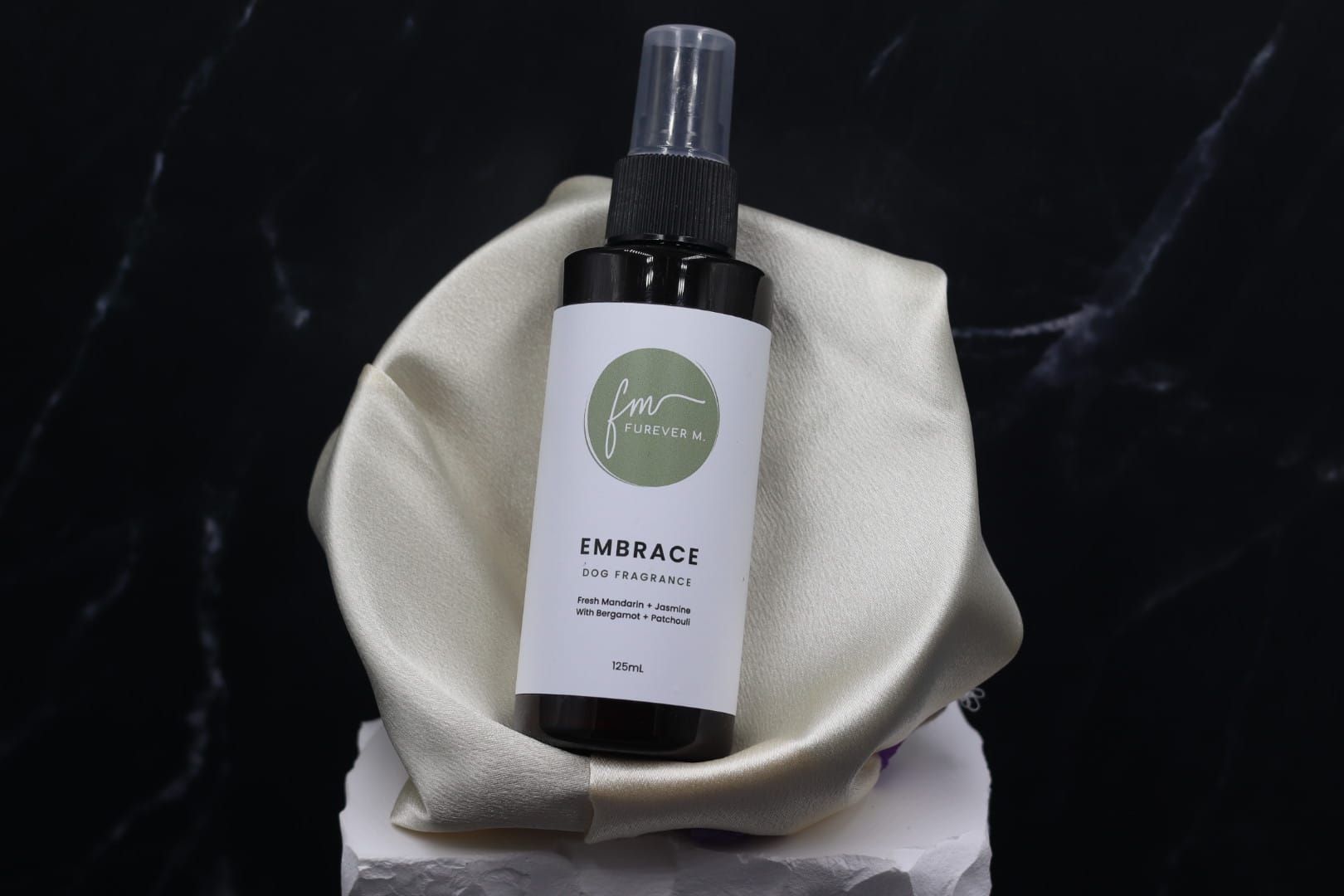 A black spray bottle of the Embrace dog cologne with it's white label with the green Furever M logo on it, placed in a silken bowl against a black marble backdrop.