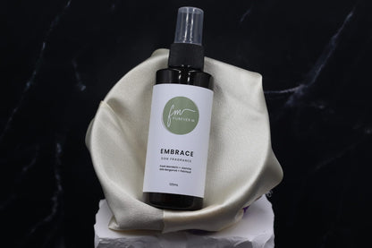 A black spray bottle of the Embrace dog cologne with it's white label with the green Furever M logo on it, placed in a silken bowl against a black marble backdrop.