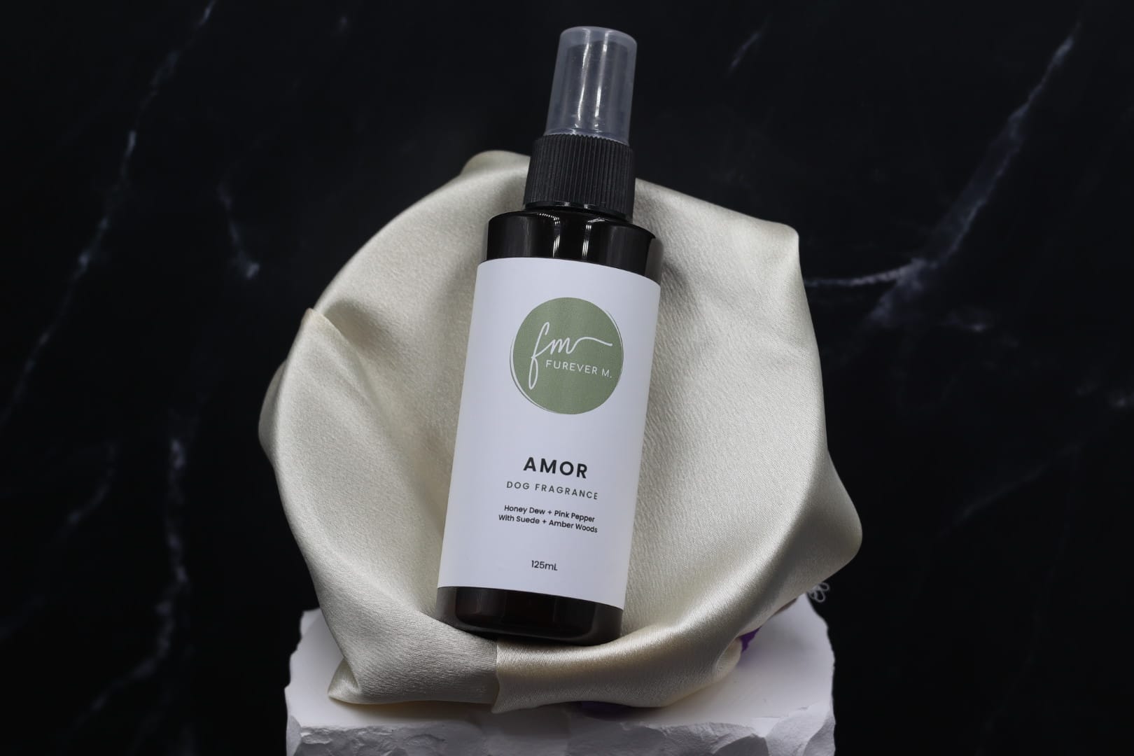 A black spray bottle of the Amor dog cologne with it's white label with the green Furever M logo on it, placed in a silken bowl against a black marble backdrop.
