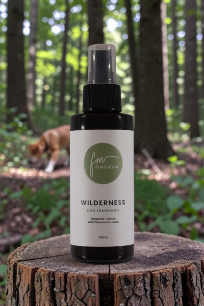 A black spray bottle of the Wilderness dog cologne with it's white label with the green Furever M logo on it, placed on a tree stump in the middle of a forest with a large brown dog in the background.