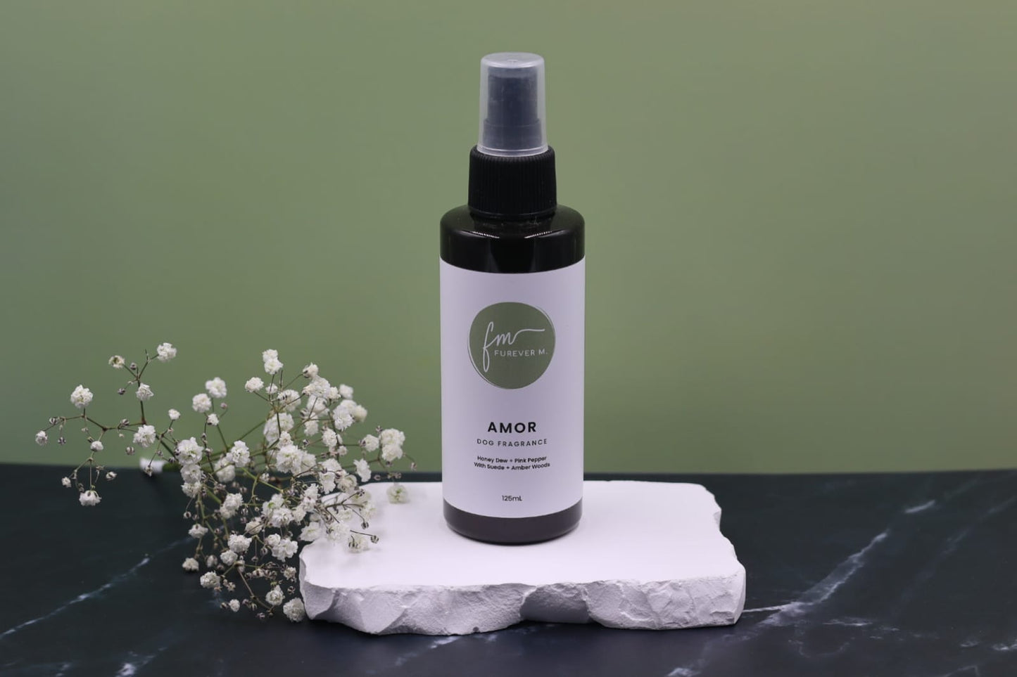 A black spray bottle of the Amor dog cologne with it's white label with the green Furever M logo on it, placed on a white marble slab placed on a black marble countertop next to a sprig of white flowers, against a sage green backdrop.