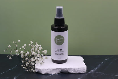 A black spray bottle of the Amor dog cologne with it's white label with the green Furever M logo on it, placed on a white marble slab placed on a black marble countertop next to a sprig of white flowers, against a sage green backdrop.