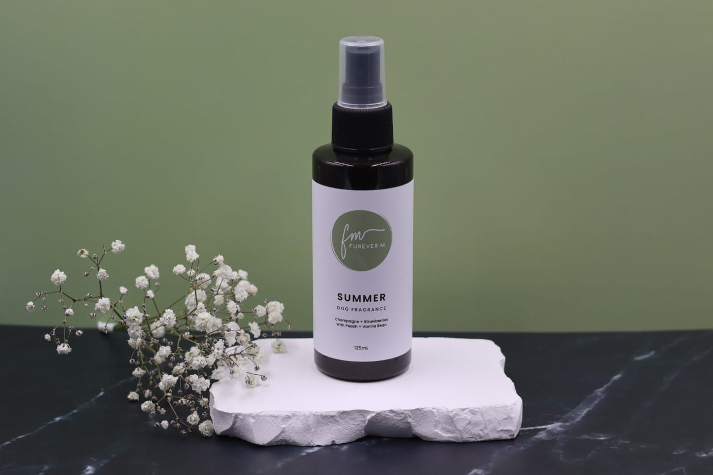 A black spray bottle of the Summer dog cologne with it's white label with the green Furever M logo on it, placed on a white marble slab placed on a black marble countertop next to a sprig of white flowers, against a sage green backdrop.