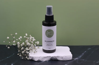 A black spray bottle of the Wilderness dog cologne with it's white label with the green Furever M logo on it, placed on a white marble slab placed on a black marble countertop next to a sprig of white flowers, against a sage green backdrop.