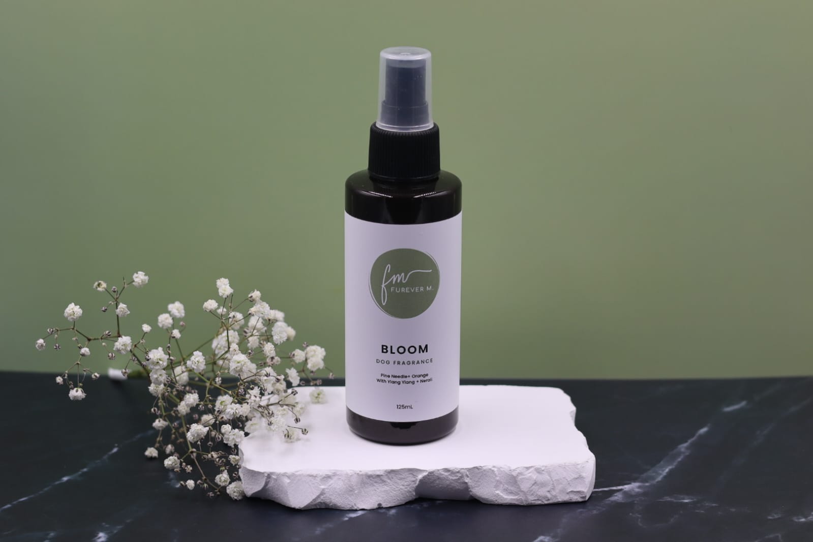 A black spray bottle of the Bloom dog cologne with it's white label with the green Furever M logo on it, placed on a white marble slab placed on a black marble countertop next to a sprig of white flowers, against a sage green backdrop.