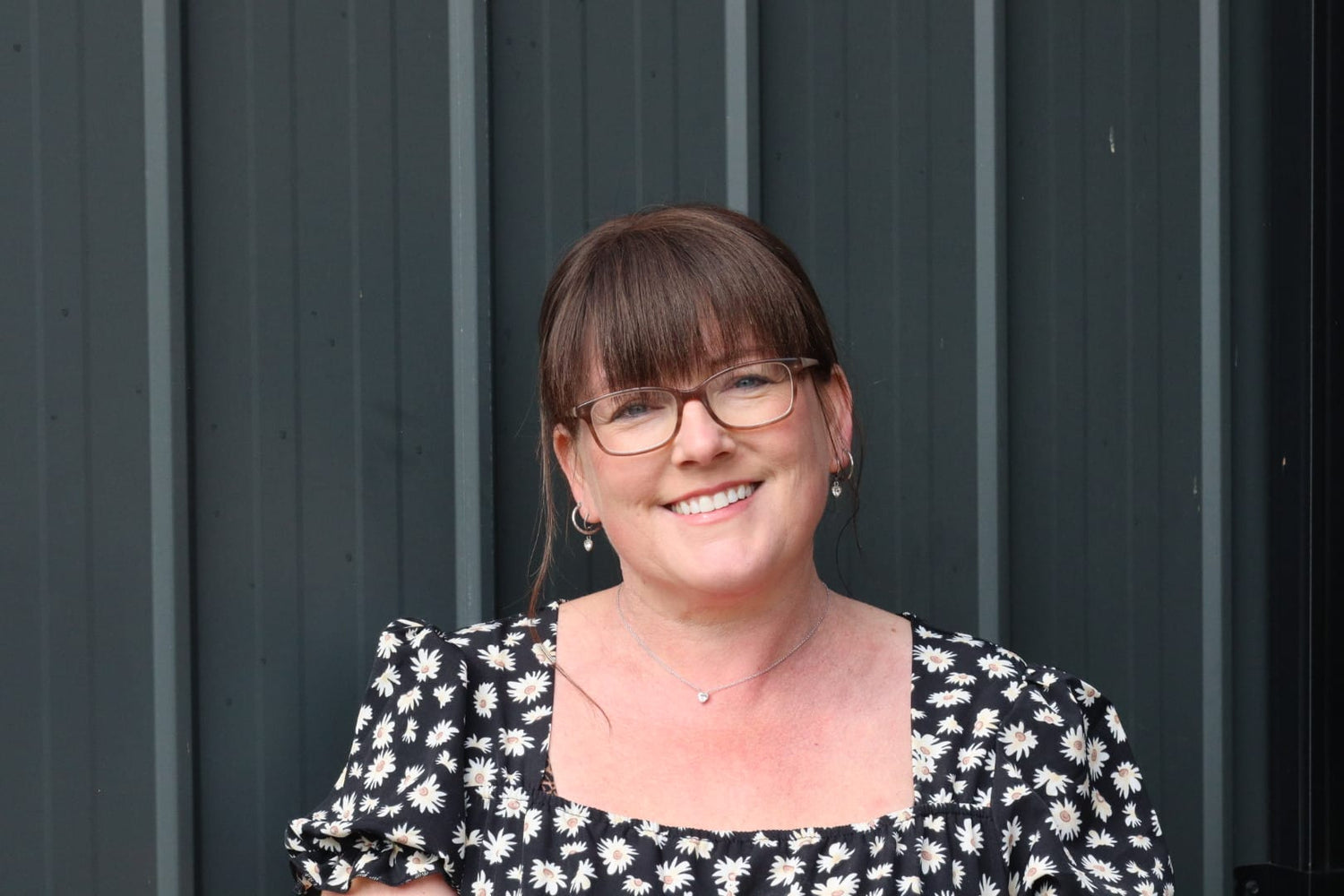The owner of Furever M, Alana, stands against her dog rooming salon smiling at the camera, wearing a black flowery dress and brown glasses.