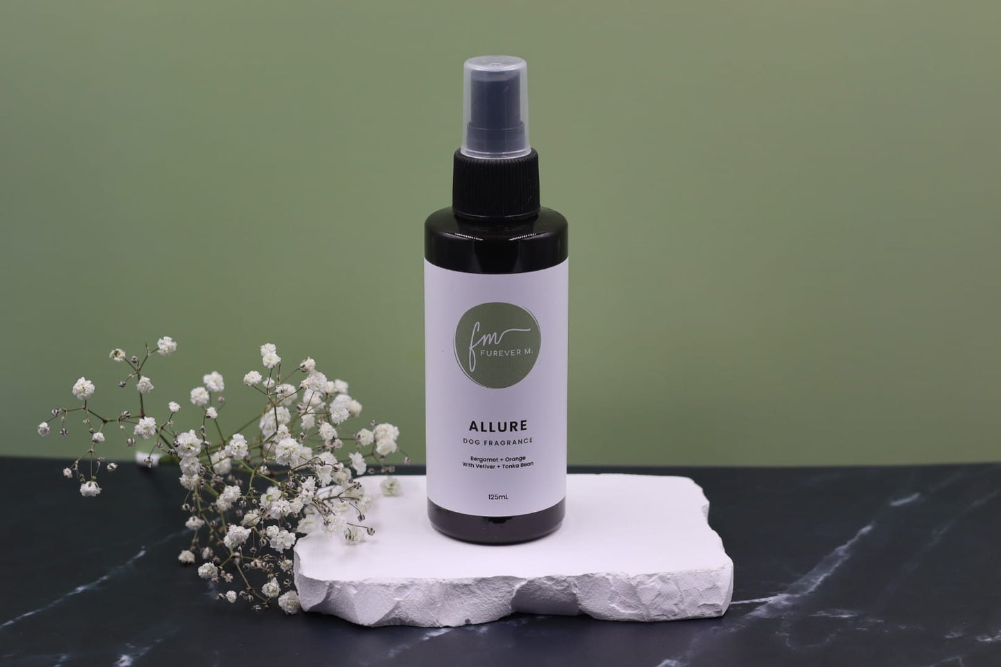 A black spray bottle of the Allure dog cologne with it's white label with the green Furever M logo on it, placed on a white marble slab placed on a black marble countertop next to a sprig of white flowers, against a sage green backdrop.